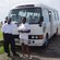 A man and a woman pose in front of a minibus with passengers on it